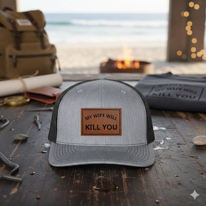 May include: Gray and black trucker hat with a brown leather patch that reads "MY WIFE WILL KILL YOU" in black text. The hat has a curved brim and mesh back. The hat is sitting on a wooden surface.