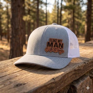 May include: Gray trucker hat with a white mesh back and a curved brim. The hat features a brown leather patch with the text "I LIKE MY MAN A Little Bit Younger" in black lettering. The hat is sitting on a wooden surface.