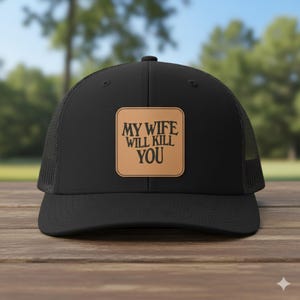 May include: Black trucker hat with a mesh back and a curved brim. The hat features a square, tan leather patch with the text "MY WIFE WILL KILL YOU" in black, bold lettering. The hat is sitting on a wooden surface.