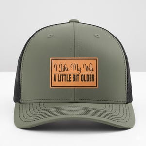 May include: Olive green trucker hat with a black mesh back. The hat features a rectangular brown leather patch with the text "I Like My Wife A Little Bit Older" in black lettering. The brim is curved and has stitched detailing.