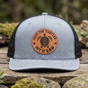 May include: Gray trucker hat with a black mesh back and a brown leather patch. The patch features a turtle design and the text "SAVE THE TURTLES HIT IT RAW". The hat has a curved brim.