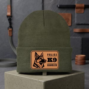 May include: An olive green knit beanie featuring a rectangular tan patch on the front. The patch displays a black graphic of a German Shepherd dog head alongside the black text that reads POLICE K9 HANDLER in a bold, block font.