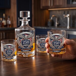 May include: A clear glass decanter and three matching whiskey glasses featuring a graphic of an eagle, the American flag, and the text United States of America 250 Years Anniversary 1776-2026 Powell. The glassware contains an amber liquid.