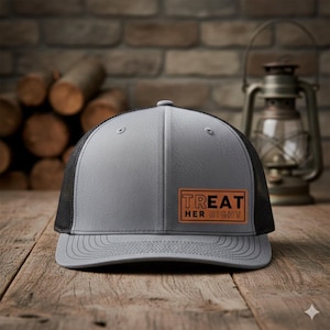 May include: A gray trucker hat with a black mesh back and a curved brim. The hat features a rectangular patch with the words "TREAT HER RIGHT" in a bold, sans-serif font. The hat is displayed on a wooden surface.