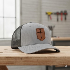 May include: Gray trucker hat with a black mesh back and a brown leather patch. The patch features a cross and nail design. The hat is sitting on a wooden surface.
