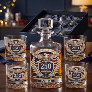 May include: A clear glass decanter and four matching whiskey glasses, each featuring a graphic of a bald eagle, the American flag, and the text United States of America 250 Years Anniversary 1776-2026 Powell. The set is presented in a dark blue box.