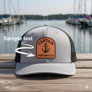 May include: A gray and black trucker hat featuring a tan leather-like patch on the front. The patch displays a black anchor graphic with the text CAPTAIN above and PERSONALIZE below. The hat includes the white text Sample text with an arrow.