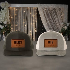 May include: Two trucker hats featuring rectangular faux leather patches. The dark gray hat displays the text HERS SINCE 2026, while the light gray hat displays the text HIS SINCE 2026. Both hats have a structured front panel and mesh back.