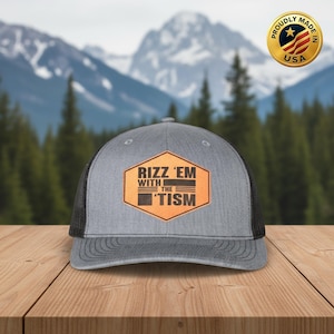 May include: Gray trucker hat with a black mesh back and a curved brim. The front features a brown hexagon patch with the words "Rizz 'Em With The 'Tism" in white and black. A gold "Proudly Made in USA" emblem is visible.