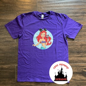 May include: Purple t-shirt with a graphic of Ariel from The Little Mermaid wearing Mickey Mouse ears and holding a Mickey Mouse ice cream bar. The graphic is in a circle with a blue background. The t-shirt has the text "Core Memory Designs" on the bottom right corner.
