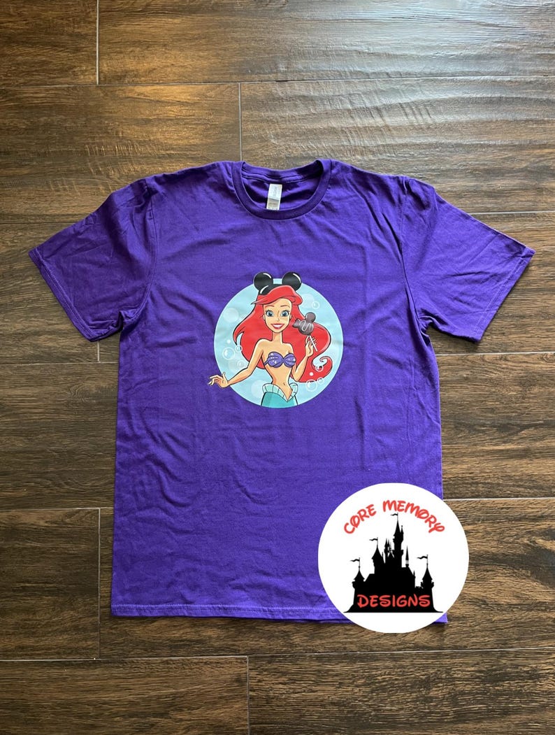 May include: Purple t-shirt with a graphic of Ariel from The Little Mermaid wearing Mickey Mouse ears and holding a Mickey Mouse ice cream bar. The graphic is in a circle with a blue background. The t-shirt has the text "Core Memory Designs" on the bottom right corner.