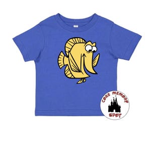 May include: A blue t-shirt featuring a yellow cartoon fish with black outlines. The fish has large eyes and a smiling expression. A circular logo with a castle silhouette and the words "CORE MEMORY SPOT" is on the bottom right.