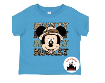 May include: A light blue t-shirt with a cartoon image of Mickey Mouse wearing a safari hat. The text "Mickey Mouse Mickey" is printed on the shirt in a brown, black, and white animal print. The shirt has a small white tag with the text "Core Memory Designs" printed on it.