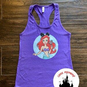 May include: Purple tank top with a graphic of Ariel from The Little Mermaid wearing Mickey Mouse ears and holding a Mickey Mouse pretzel. The graphic is in a circle with a blue background and white bubbles. The tank top has a racerback style. The text "CORE MEMORY DESIGNS" is on a white circle with a black silhouette of Cinderella's castle.