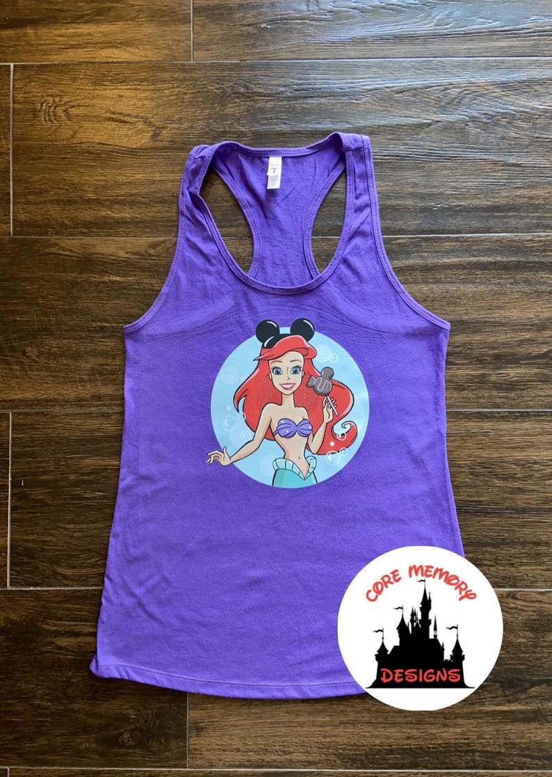May include: Purple tank top with a graphic of Ariel from The Little Mermaid wearing Mickey Mouse ears and holding a Mickey Mouse pretzel. The graphic is in a circle with a blue background and white bubbles. The tank top has a racerback style. The text "CORE MEMORY DESIGNS" is on a white circle with a black silhouette of Cinderella's castle.