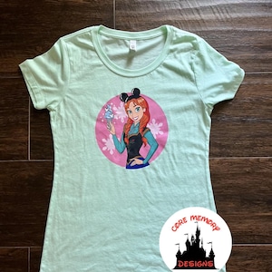 May include: A light green t-shirt featuring a cartoon illustration of a character with red hair, wearing black mouse ears, and holding an ice cream cone. The design is set within a pink circle with floral accents. The shirt has a round neckline.