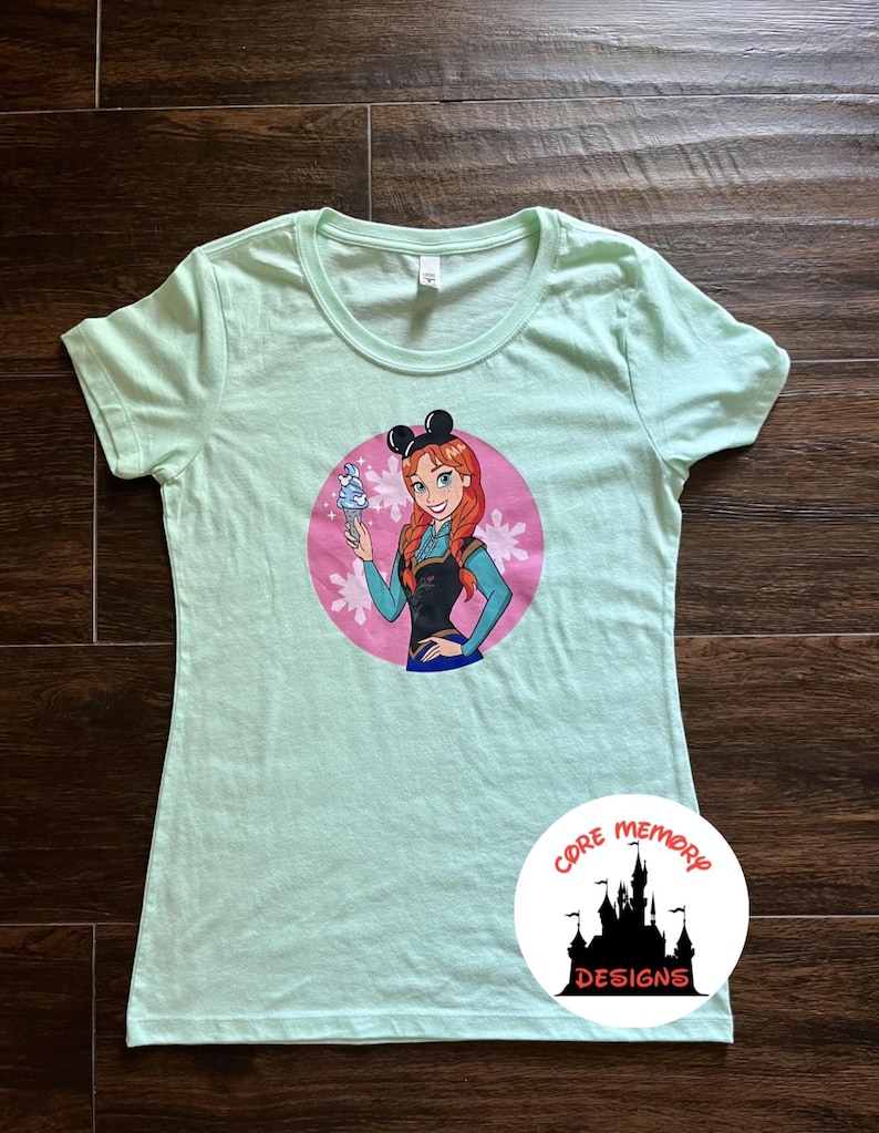 May include: A light green t-shirt featuring a cartoon illustration of a character with red hair, wearing black mouse ears, and holding an ice cream cone. The design is set within a pink circle with floral accents. The shirt has a round neckline.