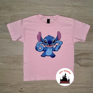 May include: A light pink t-shirt featuring a blue cartoon character holding the numbers 6 and 7. The character has large ears and a wide smile. A circular logo with text and a castle design is in the lower right corner.