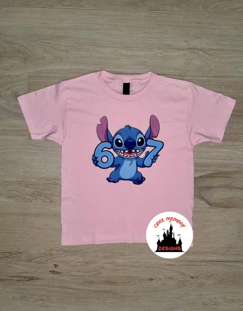 May include: A light pink t-shirt featuring a blue cartoon character holding the numbers 6 and 7. The character has large ears and a wide smile. A circular logo with text and a castle design is in the lower right corner.