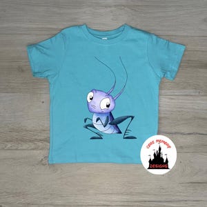 May include: A bright turquoise short-sleeved t-shirt featuring a central graphic of a purple cartoon cricket with large eyes and long antennae. A circular white logo in the bottom right corner displays a black castle silhouette and the text Core Memory Designs.