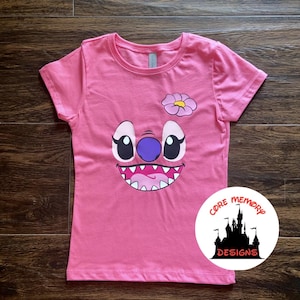 May include: Pink short-sleeved t-shirt with a cartoon Stitch face graphic. The face is purple, blue, and white with a pink flower on top. The shirt has the text "Core Memory Designs" in a circle with a black castle graphic.