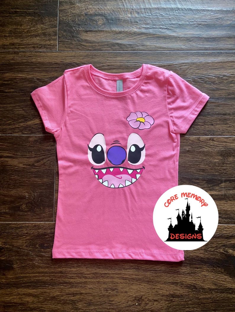 May include: Pink short-sleeved t-shirt with a cartoon Stitch face graphic. The face is purple, blue, and white with a pink flower on top. The shirt has the text "Core Memory Designs" in a circle with a black castle graphic.