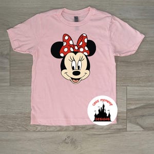 Puede incluir: Camiseta rosa claro con un dise&ntilde;o de Minnie Mouse. El dise&ntilde;o muestra la cara de Minnie, con orejas negras y un lazo rojo con lunares blancos. La camiseta tiene mangas cortas y cuello redondo.