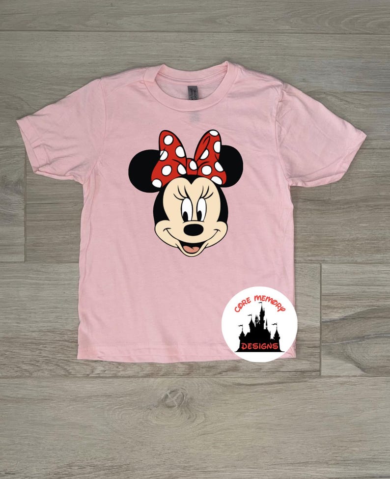 Puede incluir: Camiseta rosa claro con un dise&ntilde;o de Minnie Mouse. El dise&ntilde;o muestra la cara de Minnie, con orejas negras y un lazo rojo con lunares blancos. La camiseta tiene mangas cortas y cuello redondo.