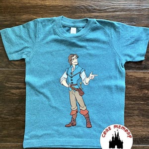 May include: Teal short-sleeved t-shirt featuring a cartoon illustration of a male character in a blue vest, white shirt, and brown pants. The character is pointing with his right hand. A logo that says "Core Memory Spot" is on the bottom right.