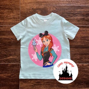 K&ouml;nnte beinhalten: Ein hellblaues T-Shirt mit einem rosa Kreis, der ein Cartoon-Bild eines M&auml;dchens mit roten Haaren zeigt, das ein Eis am Stiel h&auml;lt. Das M&auml;dchen tr&auml;gt ein blaues Kleid und eine schwarze Weste. Der Kreis ist mit wei&szlig;en Schneeflocken umgeben. Das T-Shirt hat auch ein schwarzes Mickey-Mouse-Ohren-Design auf der Oberseite. Das T-Shirt tr&auml;gt den Text "CORE MEMORY DESIGNS" in einem wei&szlig;en Kreis mit einer schwarzen Silhouette eines Schlosses.
