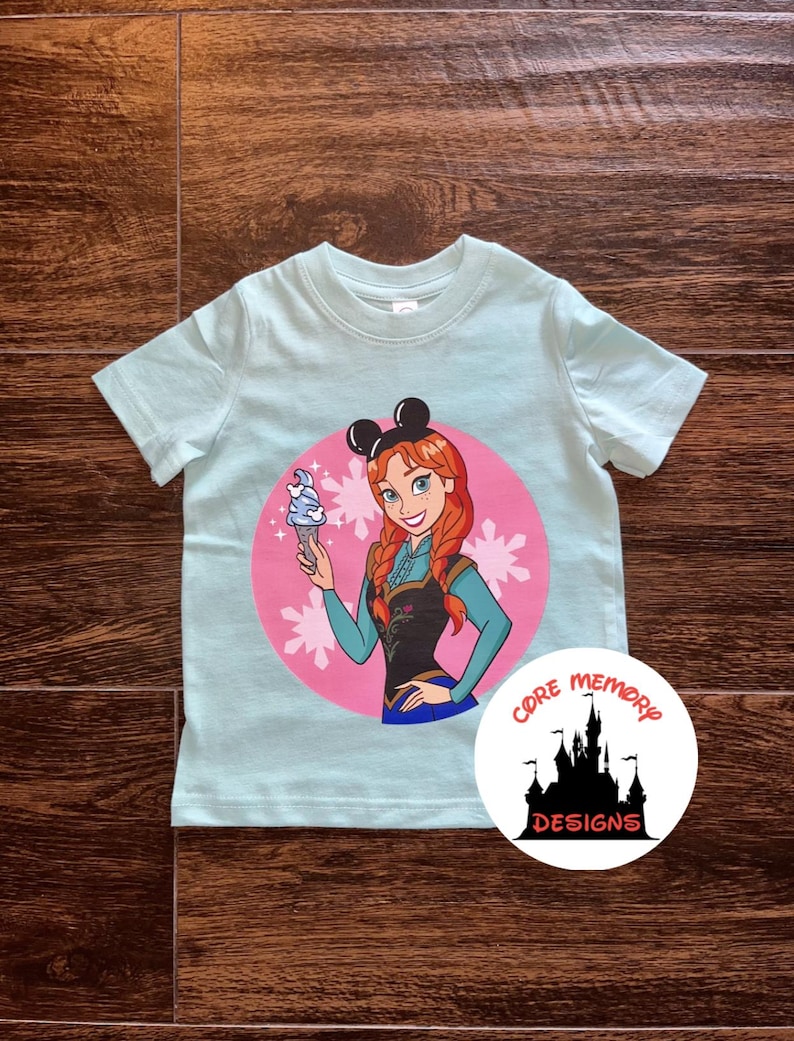 K&ouml;nnte beinhalten: Ein hellblaues T-Shirt mit einem rosa Kreis, der ein Cartoon-Bild eines M&auml;dchens mit roten Haaren zeigt, das ein Eis am Stiel h&auml;lt. Das M&auml;dchen tr&auml;gt ein blaues Kleid und eine schwarze Weste. Der Kreis ist mit wei&szlig;en Schneeflocken umgeben. Das T-Shirt hat auch ein schwarzes Mickey-Mouse-Ohren-Design auf der Oberseite. Das T-Shirt tr&auml;gt den Text "CORE MEMORY DESIGNS" in einem wei&szlig;en Kreis mit einer schwarzen Silhouette eines Schlosses.