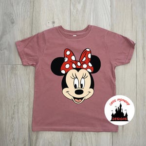 Puede incluir: Camiseta de color rosa polvoriento con un dise&ntilde;o de Minnie Mouse. El dise&ntilde;o muestra la cara de Minnie con orejas negras, un lazo rojo con lunares blancos y una expresi&oacute;n sonriente. La camiseta tiene mangas cortas y cuello redondo. El logotipo de Core Memory Designs est&aacute; en la parte inferior derecha.