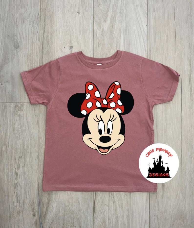 Puede incluir: Camiseta de color rosa polvoriento con un dise&ntilde;o de Minnie Mouse. El dise&ntilde;o muestra la cara de Minnie con orejas negras, un lazo rojo con lunares blancos y una expresi&oacute;n sonriente. La camiseta tiene mangas cortas y cuello redondo. El logotipo de Core Memory Designs est&aacute; en la parte inferior derecha.