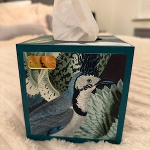 May include: A square teal tissue box cover featuring a detailed illustration of a bird with a blue crest and white plumage surrounded by green foliage. A small rectangular gold-coloured metal plaque is attached to the upper left corner of the front panel.