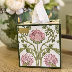 May include: A square tissue box cover featuring a white background with a repeating pattern of pink flowers and green stems. A small rectangular gold-coloured metal plaque is attached to the upper left corner, displaying the text True North Designs.