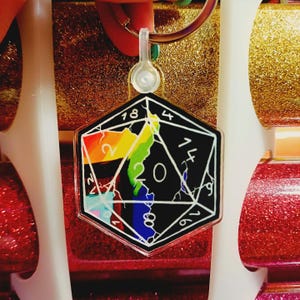 Könnte beinhalten: Ein schwarz-weißer D20-Würfel-Schlüsselanhänger mit einem Regenbogen-Design. Der Schlüsselanhänger hat die Zahlen 2, 4, 6, 8, 14, 16, 18 und 20. Der Schlüsselanhänger ist an einem silbernen Schlüsselring befestigt.