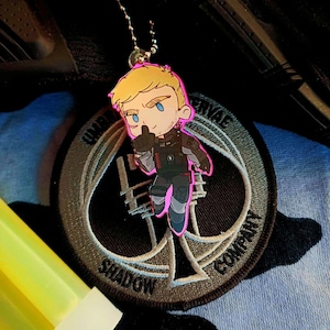 Puede incluir: Un llavero de estilo caricatura con un personaje rubio vestido con un atuendo negro y gris con detalles en rosa. El llavero está sujeto a una cadena plateada. El fondo incluye un parche con las palabras "Shadow Company" y "Umbra Servae".