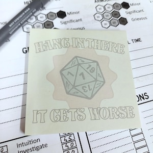 以下が含まれることがあります： 「HANG IN THERE IT GETS WORSE」の文字とサイコロが描かれたステッカー。薄い緑色で、背景は薄茶色。黒いペンとゲームシートが背景に写っています。