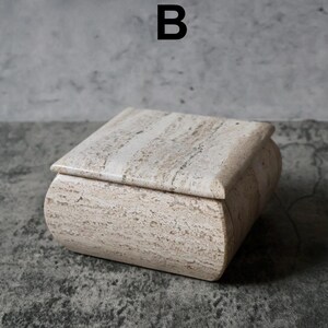 May include: A rectangular, beige stone box with a curved lid. The box has a natural, textured appearance with subtle variations in color. The letter "B" is printed in black above the box. This decorative box is suitable for storing small items.