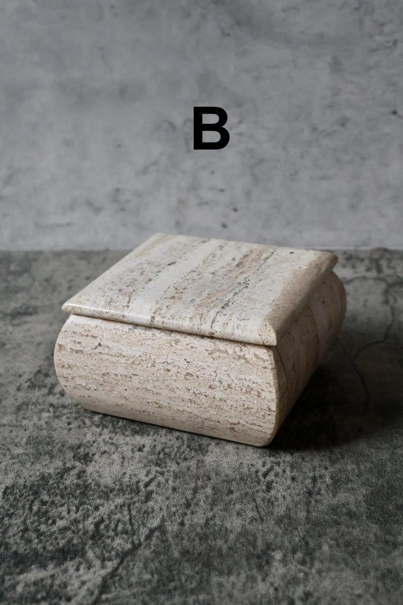 May include: A rectangular, beige stone box with a curved lid. The box has a natural, textured appearance with subtle variations in color. The letter "B" is printed in black above the box. This decorative box is suitable for storing small items.