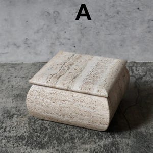 May include: A rectangular, beige stone box with a lid. The box has rounded edges and a natural, textured surface. The letter "A" is printed in black on the background.