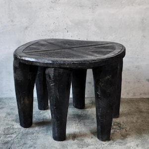 May include: A black wooden stool with a round, textured top and four thick, tapered legs. The surface of the seat features a carved, cross-like pattern. The entire piece has a dark, matte finish with visible wood grain and hand-carved details.