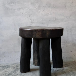 May include: A dark brown, round wooden stool featuring a thick, flat top surface and four sturdy, cylindrical legs. The wood displays a deep, charred-like finish with visible natural grain patterns and a smooth, polished texture throughout the piece.