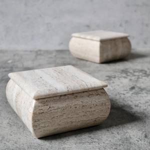 May include: Two beige stone boxes with rounded edges and square lids are displayed on a gray surface. The boxes have a natural, textured appearance, with subtle variations in color and pattern. The boxes are likely used for storage or decorative purposes.