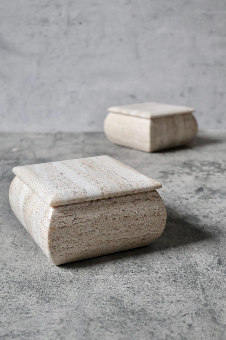 May include: Two beige stone boxes with rounded edges and square lids are displayed on a gray surface. The boxes have a natural, textured appearance, with subtle variations in color and pattern. The boxes are likely used for storage or decorative purposes.
