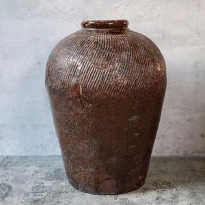 May include: A large, antique-style brown ceramic jar with a textured, ridged surface. The jar has a rounded body and a narrow neck, suggesting it could be used for storage or decoration. The color is a deep, earthy brown.