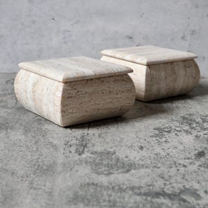 May include: Two beige stone boxes with rounded sides and flat, square lids. The boxes have a natural, textured appearance, and are set against a mottled gray background. The boxes are likely for storage or decorative purposes.