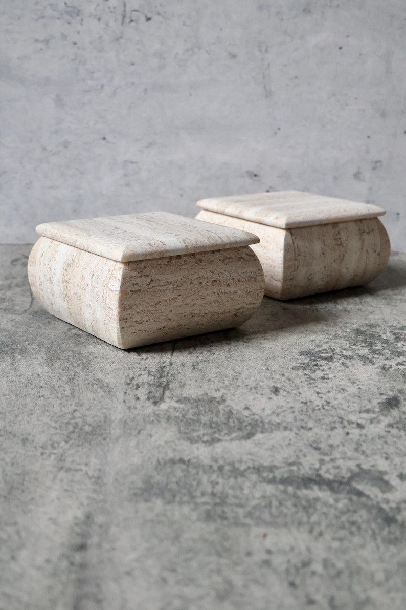 May include: Two beige stone boxes with rounded sides and flat, square lids. The boxes have a natural, textured appearance, and are set against a mottled gray background. The boxes are likely for storage or decorative purposes.