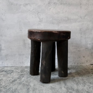 May include: A dark brown wooden stool with a thick, circular seat and three sturdy, cylindrical legs. The wood features a visible, weathered texture with natural variations in color and tone, showcasing a raw, hand-carved appearance throughout.