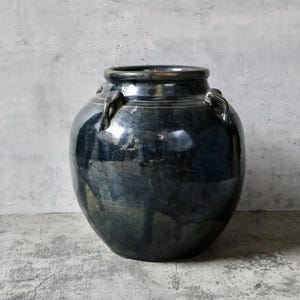May include: A dark blue glazed ceramic jar with a rounded body and a wide opening. The jar has two small handles on the sides and a glossy finish. The background is a light gray concrete.